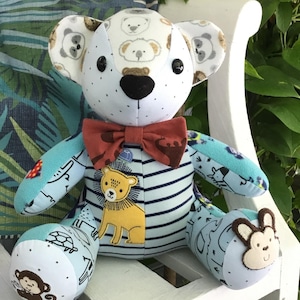 May include: A blue and white patchwork teddy bear with a red bow tie. The bear is sitting on a white bench and has a variety of animal designs on its fabric, including monkeys, lions, and turtles.
