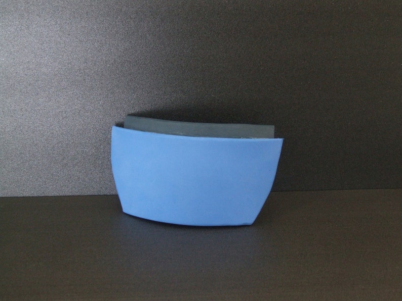 Op de afbeelding: Een gebogen schuimobject in blauw en zwart. Het object bestaat uit twee delen schuim, een blauw en een zwart. Het blauwe schuim bevindt zich aan de onderkant en het zwarte schuim aan de bovenkant.