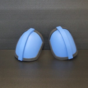 Op de afbeelding: Een paar blauwe schuimschouderpads met zwarte trim. De pads hebben een gewelfde vorm en een zwarte band die in het midden loopt.