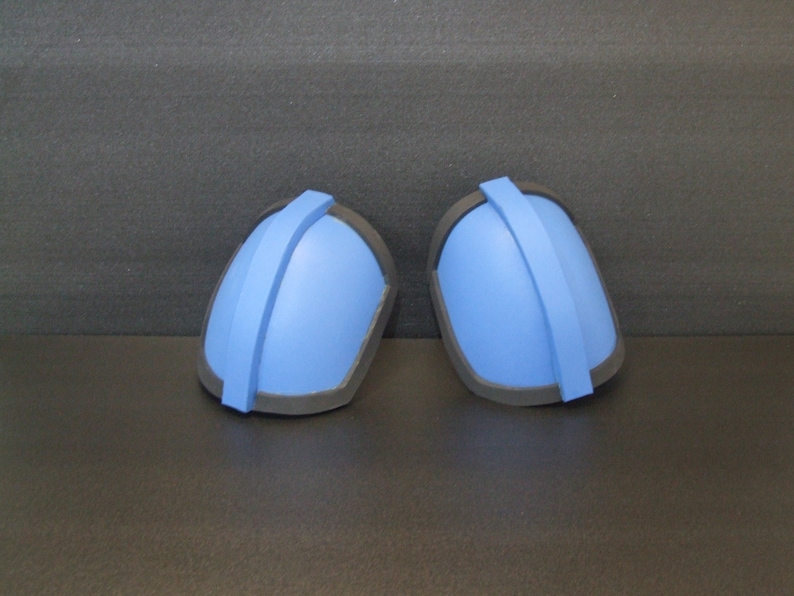Op de afbeelding: Een paar blauwe schuimschouderpads met zwarte trim. De pads hebben een gewelfde vorm en een zwarte band die in het midden loopt.