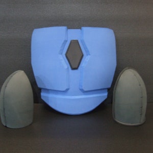 Puede incluir: Una pieza de pecho de espuma azul con dos hombreras de espuma negra. La pieza de pecho tiene un diseño en forma de diamante elevado con un centro negro.