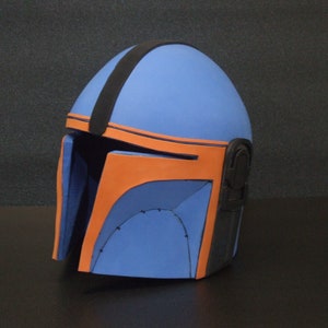 Può includere: Un casco blu e arancione con una striscia nera e una visiera nera. Il casco è fatto di schiuma e ha una superficie testurizzata.