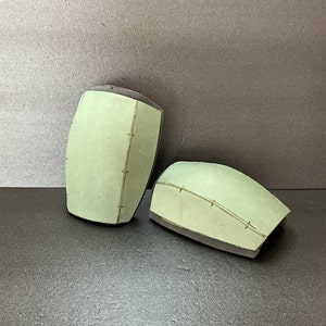 May include: Two ceramic bowls with a light green glaze and a dark brown rim. The bowls have a stitched design on the surface.