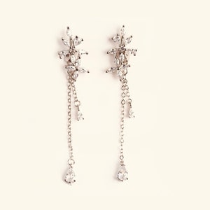 Può includere: Un paio di orecchini pendenti in argento con un gruppo di pietre di zircone cubico trasparenti nella parte superiore e un'unica pietra a forma di lacrima nella parte inferiore di ogni orecchino.