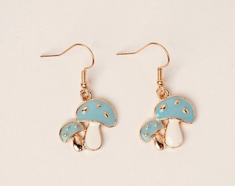 May include: A pair of gold-colored mushroom earrings. Each earring features two mushrooms with light blue caps, white stems, and gold accents. The earrings have a hook closure.