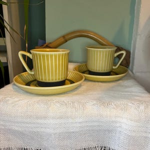 Puede incluir: Dos tazas de café de cerámica amarilla con ribetes negros y un patrón a rayas sobre un mantel blanco con flecos. Las tazas están sobre platillos a juego.