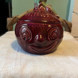 Könnte beinhalten: Ein keramikbehälter in Form einer Rote Bete mit Deckel. Der Behälter hat eine tiefrote Farbe mit einem lächelnden Gesichtsdesign und dem Wort "BEETROOT" auf dem Deckel. Die Oberseite des Behälters hat grüne und rote Akzente.