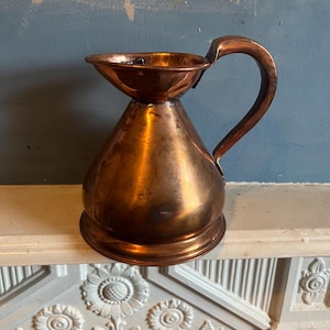 May include: A copper pitcher with a handle. The pitcher has a wide, flared top and a rounded body that tapers to a smaller base. The handle is curved and attached to the side of the pitcher. The copper has a warm, reddish-brown color.