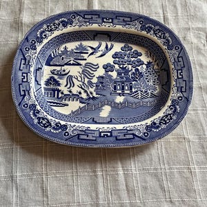 May include: An oval ceramic platter featuring a blue and white Willow pattern. The design depicts a landscape with a pagoda, a bridge, willow trees, and two birds in flight. A ruler at the bottom shows the platter is approximately 12 inches wide.