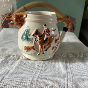 Puede incluir: Una jarra de cerámica de color crema con un pico en forma de cabeza de zorro y un asa. El exterior está decorado con un relieve colorido que muestra a un jinete sobre un caballo marrón acompañado de perros de caza en un campo de hierba.