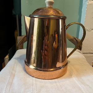 May include: A vintage copper and brass coffee pot with a lid and a white ceramic knob. The pot has a curved handle and a spout for pouring. The base and the lid are copper, while the body is brass.