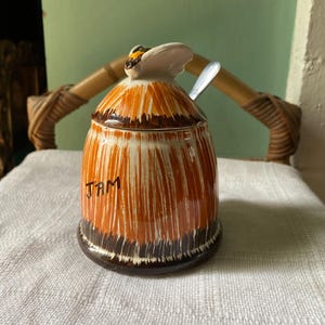 May include: A ceramic jam jar with a bee-shaped lid and a small spoon. The jar is cylindrical with a brown and orange striped pattern and the word "JAM" written on the front. The jar sits on a white cloth.