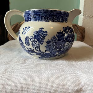 Puede incluir: Una jarra pequeña y redonda de color crema con un patrón azul de Willow. La jarra tiene un asa y un pico. El patrón azul representa un paisaje con edificios, árboles y figuras. La jarra está sobre una tela blanca.