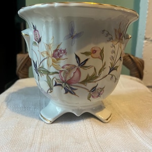 Puede incluir: Una maceta de cerámica blanca con borde dorado y tres patas. La maceta está decorada con un diseño floral y de mariposas en rosa, amarillo y azul. La maceta tiene dos asas.