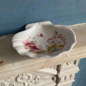 Puede incluir: Un plato de porcelana blanca en forma de concha con un diseño floral en tonos de rosa, rojo, amarillo y azul. El plato presenta una mariposa en el centro.