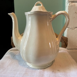 May include: Cream-coloured ceramic teapot with a lid and curved handle. The teapot has a spout and sits on a small base. The surface has a crackle glaze effect. The teapot is likely vintage or antique.