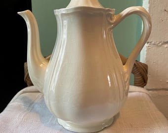 May include: Cream-colored ceramic teapot with a lid and curved handle. The teapot has a spout and sits on a small base. The surface has a crackle glaze effect. The teapot is likely vintage or antique.