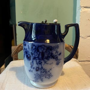 May include: A vintage blue and white porcelain jug with a floral pattern. The jug has a dark blue top and handle, with a white base. The floral pattern is in shades of blue. The jug has a lid with a white knob.