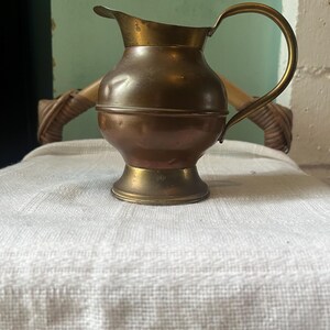 May include: A vintage copper and brass jug with a curved handle and a spout. The jug has a bulbous body and a flared base. The metal has a warm, aged patina. The jug is sitting on a white, fringed cloth.