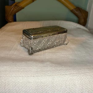 May include: A clear glass box with a silver lid. The box has a diamond pattern and is sitting on a white fabric surface.