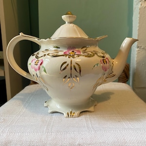 Puede incluir: Una tetera blanca con un diseño floral dorado y rosa. La tetera tiene una tapa con un pomo dorado y una base festoneada.