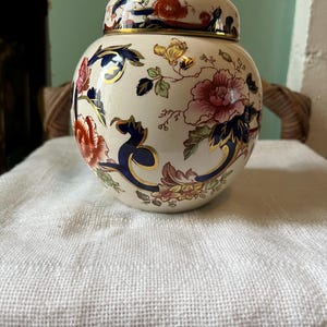 May include: A cream-coloured ceramic jar with a lid, decorated with floral patterns in red, pink, blue, and gold. The jar has a rounded shape and a gold rim, suggesting a decorative or storage item. The floral design adds a touch of elegance.