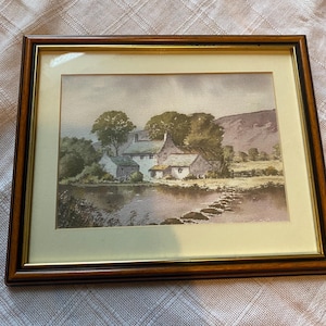 May include: A framed watercolor painting depicting a rural scene with a small village nestled near a body of water. The painting features a cluster of houses with green roofs and a stone bridge leading across the water. The sky is a soft blue with wispy clouds, and the surrounding landscape is lush and green.
