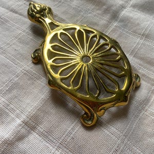 Puede incluir: Un posavasos de metal dorado en forma de tortuga con un diseño decorativo calado. El posavasos tiene un centro circular con un pequeño agujero.