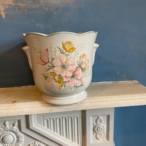 May include: White ceramic planter with a scalloped rim and two handles. The planter features a floral design with pink, yellow, and green flowers.