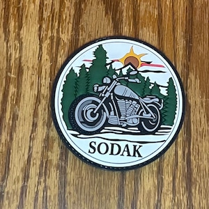 May include: A white, round patch with a black border. The patch features a black and white motorcycle with a green forest background and a yellow sun in the sky. The text "SODAK" is printed in black at the bottom of the patch.