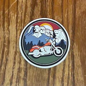 May include: A round patch with a white background featuring a red motorcycle with a mountain range and a depiction of Mount Rushmore in the background. The patch has a green border.