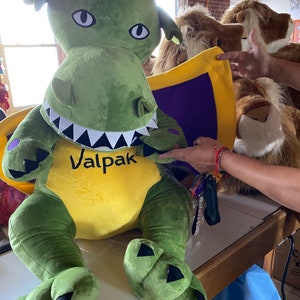 Puede incluir: Un gran dragón de peluche verde con alas amarillas y moradas. El dragón tiene una sonrisa de dientes y la palabra "Valpak" impresa en su pecho.
