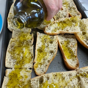 May include: A close-up of a hand pouring olive oil over slices of bread on a baking sheet. The bread is topped with black pepper.