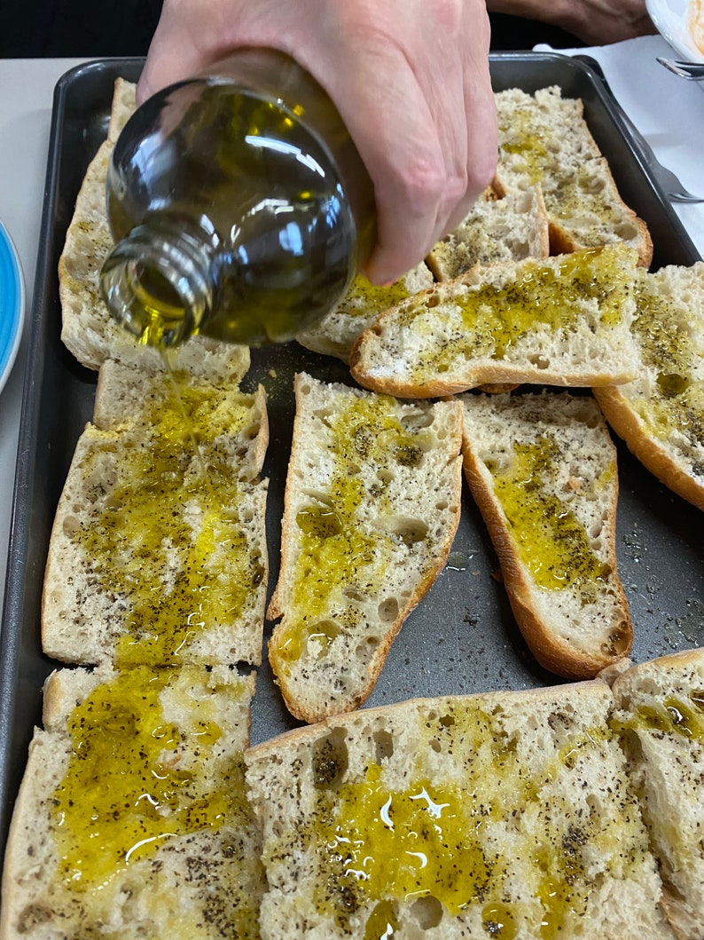 May include: A close-up of a hand pouring olive oil over slices of bread on a baking sheet. The bread is topped with black pepper.