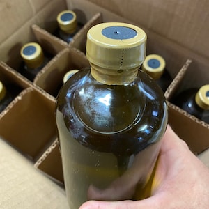 Puede incluir: Primer plano de una botella de vidrio marr&oacute;n con una tapa dorada. La botella est&aacute; en una caja de cart&oacute;n con otras botellas.