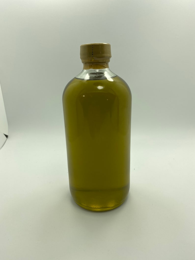 Puede incluir: Una botella de vidrio transparente con una tapa dorada llena de un l&iacute;quido verde. La botella est&aacute; sobre una superficie blanca.