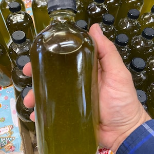 Puede incluir: Primer plano de una botella de vidrio transparente llena de un l&iacute;quido verde. La botella est&aacute; siendo sostenida por una mano, y hay muchas otras botellas similares en el fondo.