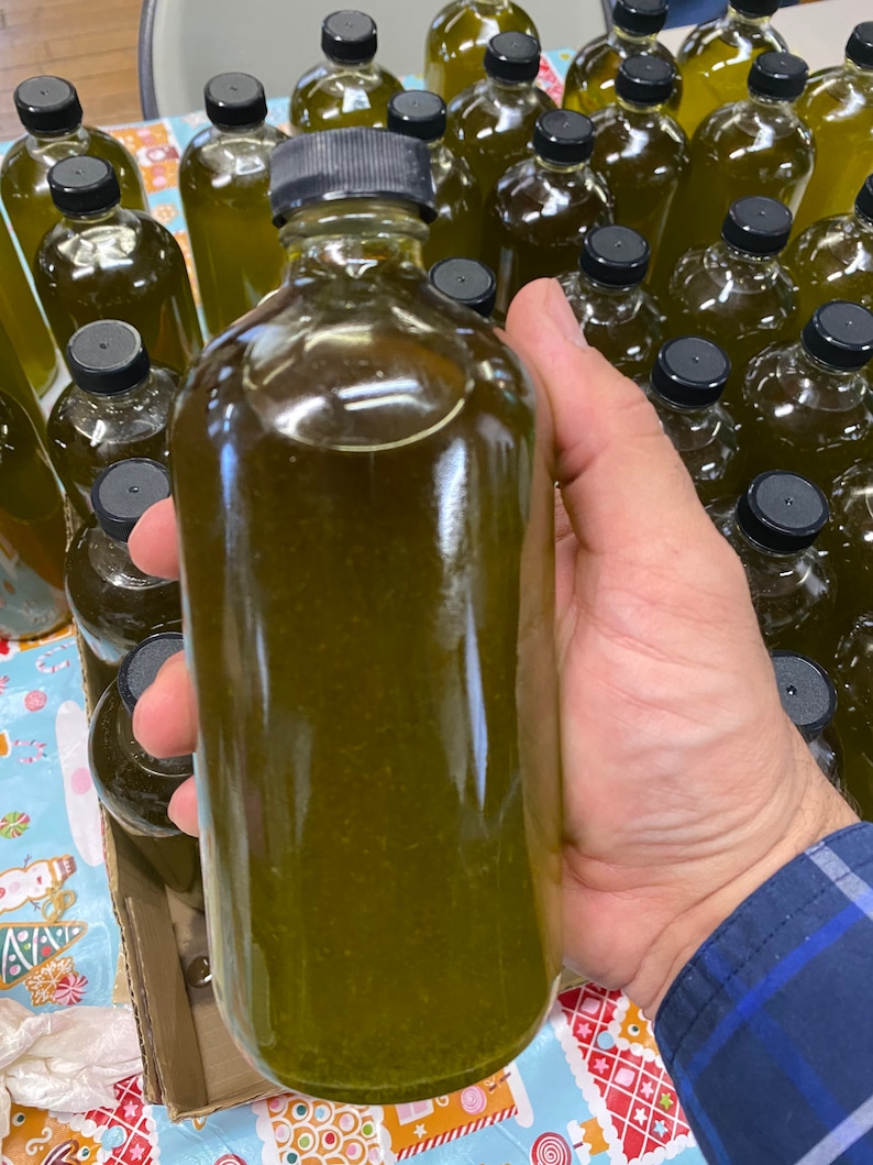 Puede incluir: Primer plano de una botella de vidrio transparente llena de un l&iacute;quido verde. La botella est&aacute; siendo sostenida por una mano, y hay muchas otras botellas similares en el fondo.