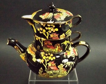 May include: A black and gold teapot with a floral design. The teapot is stacked on top of two cups, also with floral designs. The teapot and cups are on a clear acrylic stand.