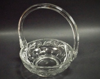 May include: Clear glass basket with a curved handle. The basket has a round bowl shape with floral etched detailing around the rim. The handle is attached to the sides of the bowl. The basket is on a dark surface.