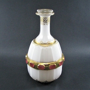 Puede incluir: Una decantador de vidrio blanco con detalles dorados y un tapón de vidrio rojo. El decantador tiene un cuerpo redondo con una superficie texturizada y un cuello largo y delgado.