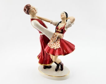 May include: A porcelain figurine depicting two figures in a dynamic dancing pose. The figures wear red and white outfits with gold trim. The male figure has brown hair, and the female figure has black hair in a braid. They stand on an oval base.