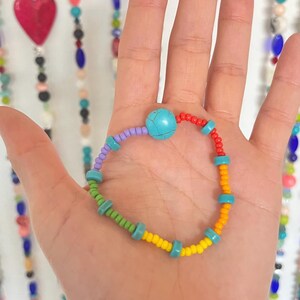 May include: A colorful beaded bracelet with a turquoise central bead. The bracelet features a variety of small beads in green, yellow, orange, red, purple, and turquoise. The bracelet is held in a hand.