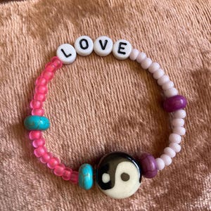May include: A pink, white, and purple beaded bracelet with the word "LOVE" spelled out in white beads. The bracelet also features a turquoise and a black and white yin yang bead.
