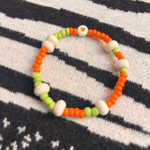 May include: A colorful beaded bracelet with orange, green, and white beads. A white bead with a small orange heart is in the center of the bracelet.