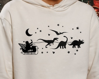 以下が含まれることがあります： 背景に三日月と星が描かれた、白いフーディー。黒いシルエットで、サンタクロースが恐竜に引かれたそりで空を飛んでいるデザインです。