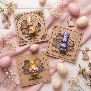 Könnte beinhalten: Osterarrangement mit drei Kraftpapierkarten mit Korb- und Eierillustrationen, Schokoladenhasen und -küken sowie dekorativen Eiern. Die Karten haben deutschen Text. Rosa Stoff, Holzhasen und Blumen ergänzen die Szene.