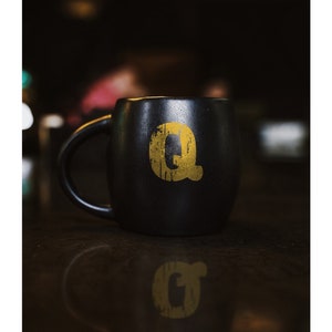 May include: A black ceramic mug with a gold letter "Q" on the side. The mug is sitting on a dark surface.
