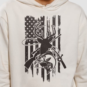 May include: A cream-colored hoodie with a black and white graphic print of a deer, fish, and hunting rifle superimposed on an American flag.