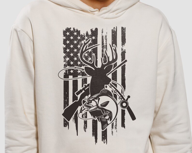 May include: A cream-colored hoodie with a black and white graphic print of a deer, fish, and hunting rifle superimposed on an American flag.